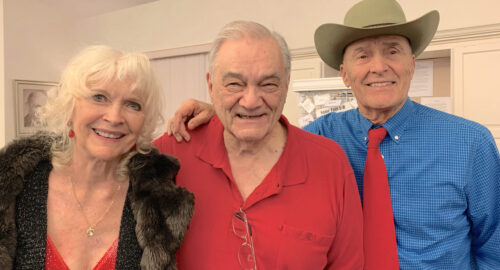 three friends standing together, woman on left in red dress, man in middle in red shirt, man on right in blue shirt and red tie and cowboy hat